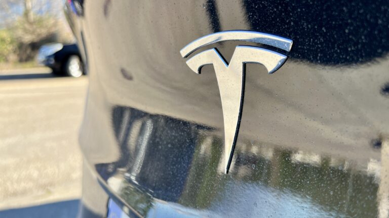 A detailed close-up view of the iconic Tesla emblem on a car's hood, highlighted by water droplets and a reflective surface.